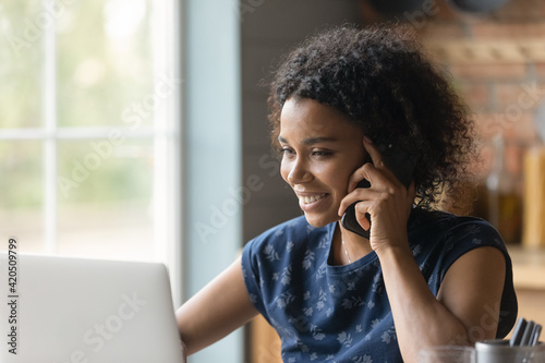 Modern way of communication. Smiling biracial female hold cell at ear talk look at pc screen. Busy young black lady advisor lawyer give phone consultation from home discuss electronic document details