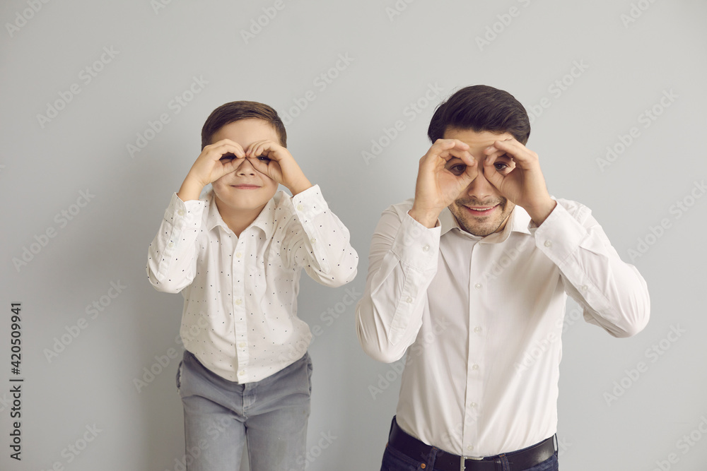 Cheerful father and little son look into imaginary binoculars made of their hands. Smiling family making imitation binoculars with fingers and looking at camera while standing on gray background.