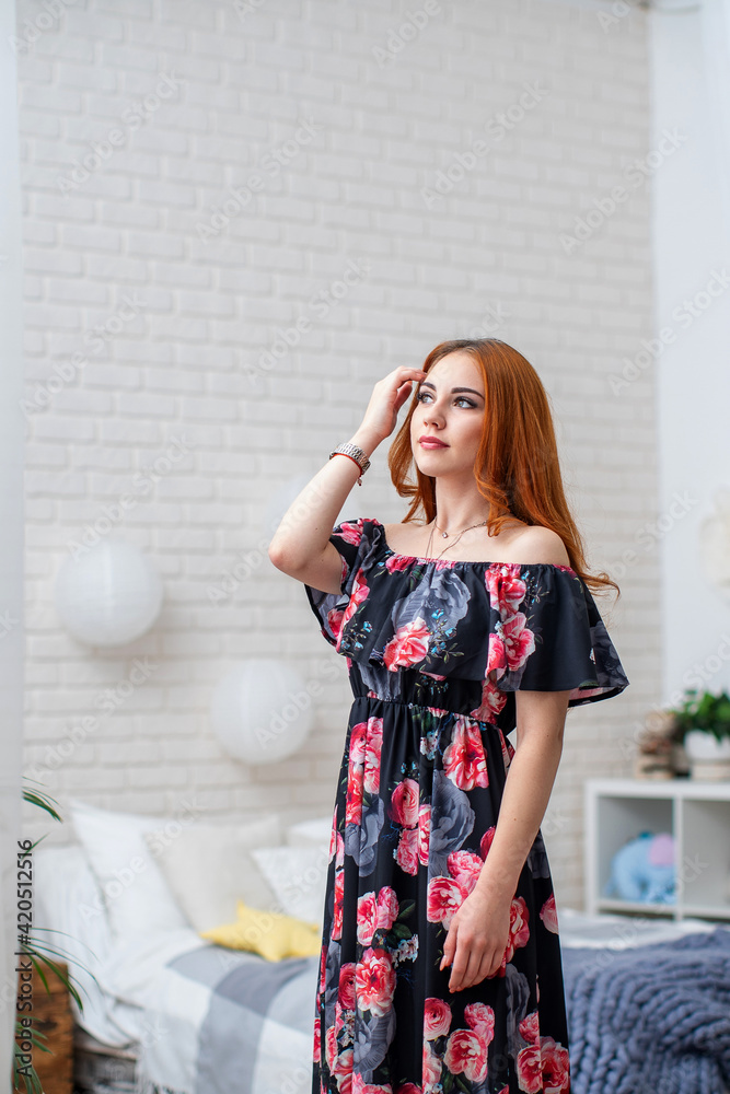 Attractive young woman in floral print dress stands in front large mirror in graceful floral interior. pretty girl with curly red hair, in beautiful dress, in floral interior, smiles. spring festival