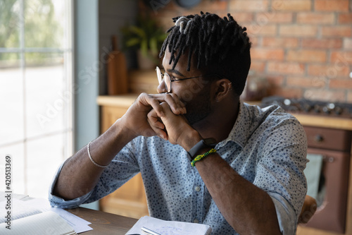 Thinking by window. Pensive afro caribbean guy student hipster sit at table leaning on folded hands reflect on training task. Concentrated young black man freelancer creating idea planning future work photo
