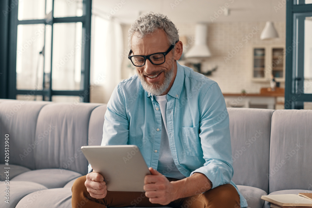 Cheerful senior man in casual clothing using digital tablet while sitting on the sofa at home Cheerful senior man in casual clothing using digital tablet while sitting on the sofa at home