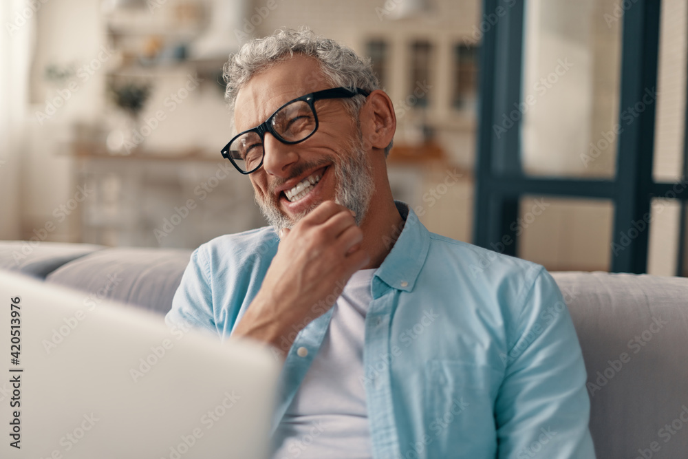 Cheerful senior man in casual clothing and eyeglasses using laptop and smiling while sitting on the sofa at home - obrazy, fototapety, plakaty 