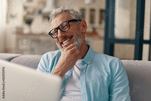 Cheerful senior man in casual clothing and eyeglasses using laptop and smiling while sitting on the sofa at home photo