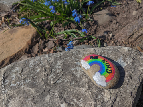 Sign be kind and a rainbow on a painted rock. Selective focus, blurred spring blue flowers in the background. Kindness, hope, love, tolerance, anti racism, inclusivness concept. Space for copy.