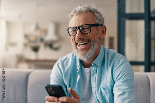 Cheerful senior man in casual clothing and eyeglasses using smart phone while sitting on the sofa at home photo
