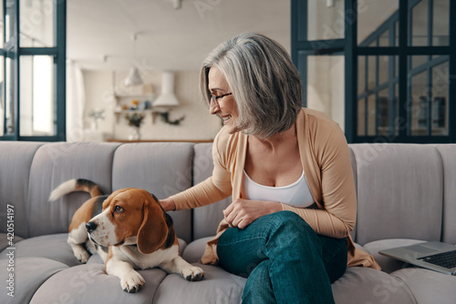 Cheerful senior woman in casual clothing spending time with her dog while sitting on the sofa at home