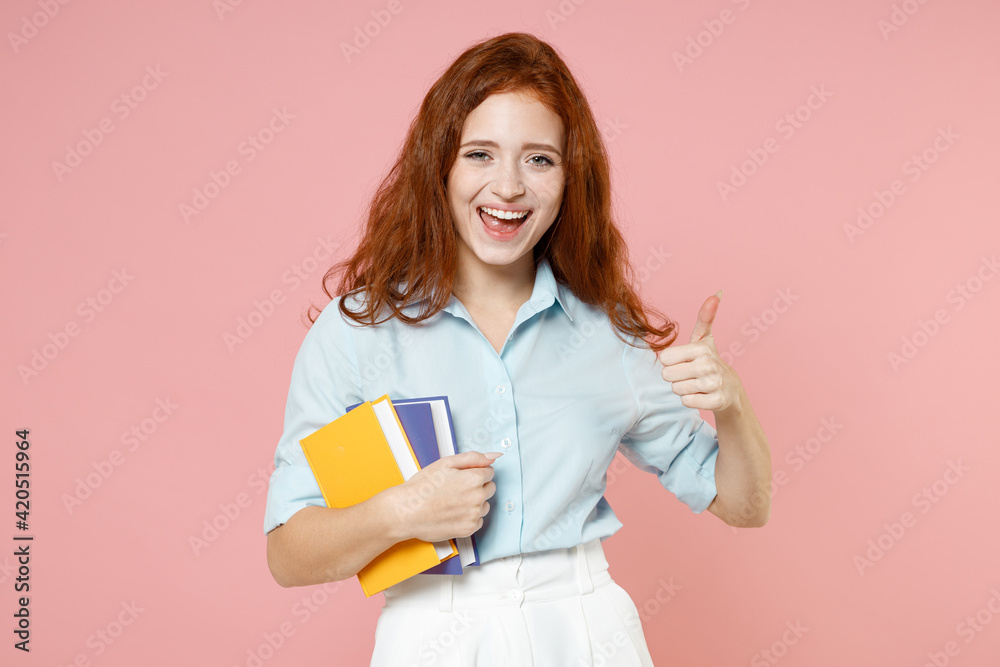 Young smiling redhead student woman in blue shirt hold book notebook show thumb up like gesture isolated on pastel pink background studio portrait. Education high school university college concept Young smiling redhead student woman in blue shirt hold book notebook show thumb up like gesture isolated on pastel pink background studio portrait. Education high school university college concept