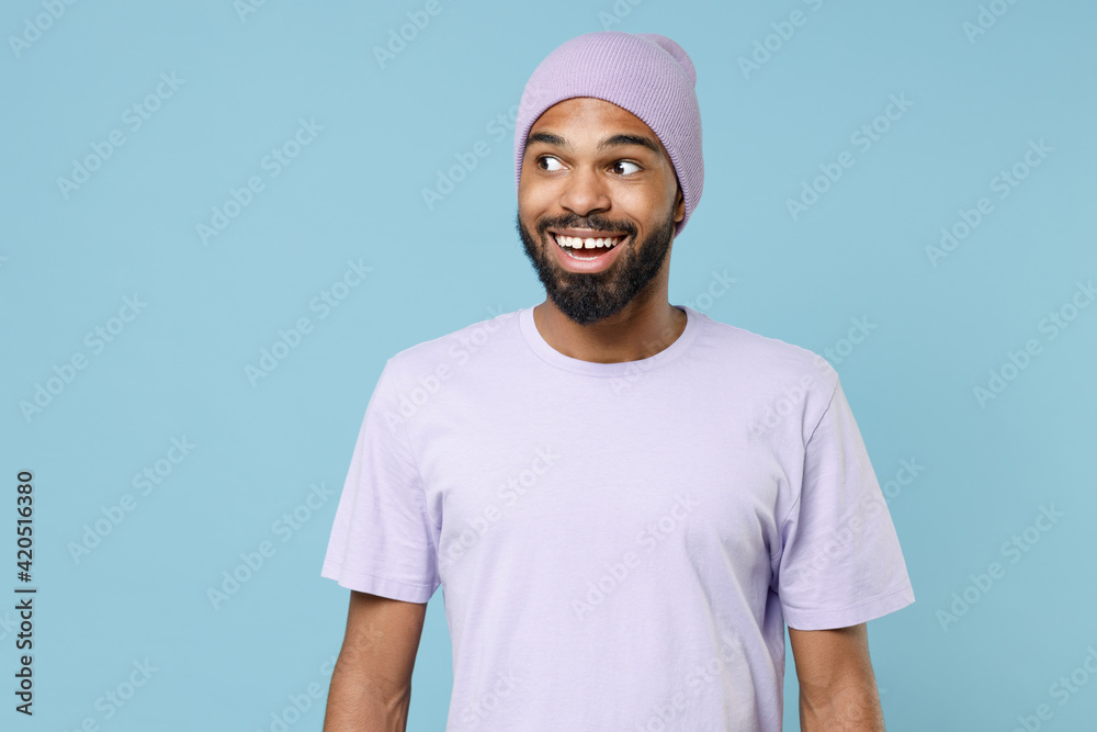 Young smiling dreamful impressed surprised student fun unshaven black dark-skinned african man 20s in violet t-shirt purple hat looking aside isolated on pastel blue color background studio portrait Young smiling dreamful impressed surprised student fun unshaven black dark-skinned african man 20s in violet t-shirt purple hat looking aside isolated on pastel blue color background studio portrait