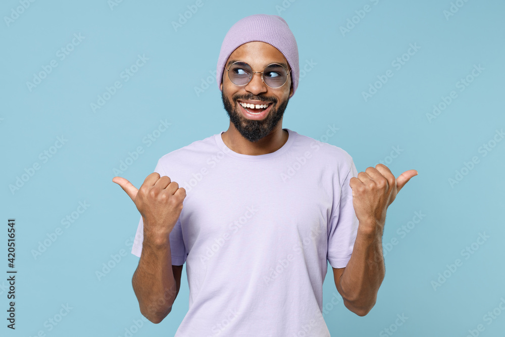 Naklejka premium Young smiling unshaven black dark-skinned african american man 20s in violet t-shirt hat glasses point thumb fingers aside on workspace area mock up isolated on pastel blue background studio portrait.