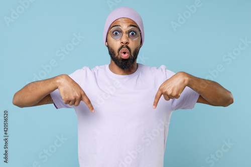 Young surprised unshaven black dark-skinned african american man 20s in violet t-shirt hat glasses point index finger down on workspace area mock up isolated on pastel blue background studio portrait.
