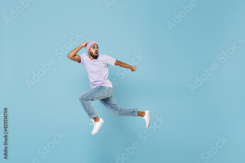 Full length of young shocked scared frightened unshaven black african man 20s in violet t-shirt hat glasses jump high run away fast hurrying isolated on pastel blue color background studio portrait.