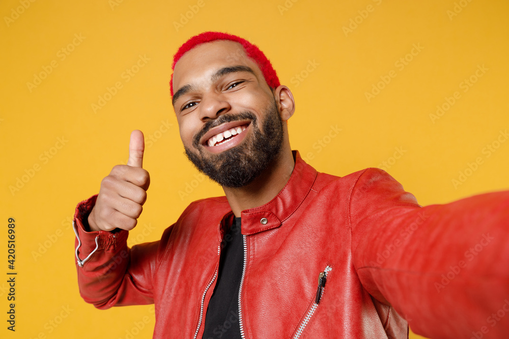 Close up young african smiling man with funky trendy pink hairdo in red leather jacket do selfie shot on mobile phone show thumb up like gesture isolated on yellow orange background studio portrait. Close up young african smiling man with funky trendy pink hairdo in red leather jacket do selfie shot on mobile phone show thumb up like gesture isolated on yellow orange background studio portrait.
