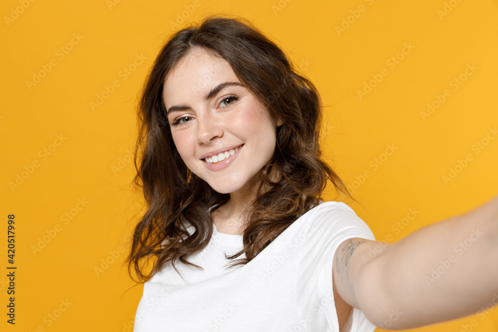 Close up young smiling european friendly happy cool caucasian woman 20s wearing white basic casual t-shirt doing selfie shot on mobile phone isolated on yellow orange color background studio portrait. Close up young smiling european friendly happy cool caucasian woman 20s wearing white basic casual t-shirt doing selfie shot on mobile phone isolated on yellow orange color background studio portrait.