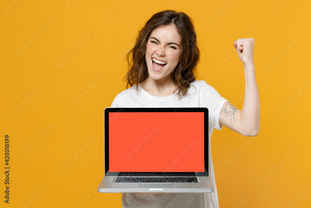 Naklejka premium Young overjoyed freelancer student woman in white basic t-shirt hold laptop pc computer with blank screen workspace area doo winner gesture clench fist celebrate isolated on yellow background studio.