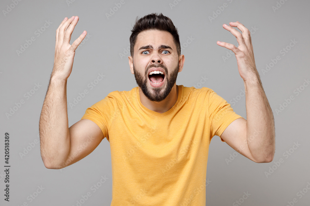 Young caucasian strict bearded handsome angry indignant expressive man 20s in casual yellow basic t-shirt spreading hands screaming shouting looking camera isolated on grey background studio portrait. Young caucasian strict bearded handsome angry indignant expressive man 20s in casual yellow basic t-shirt spreading hands screaming shouting looking camera isolated on grey background studio portrait.
