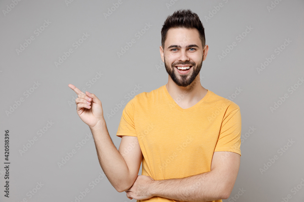 Young caucasian bearded smiling man 20s in yellow basic t-shirt point index finger aside on workspace area copy space mock up isolated on grey background studio portrait. People lifestyle concept.