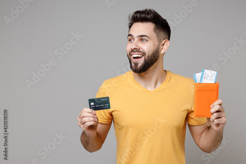 Traveler young happy tourist man 20s wearing yellow t-shirt holding passport tickets credit bank card look aside isolated on grey background Passenger travel abroad weekends getaway Journey concept.