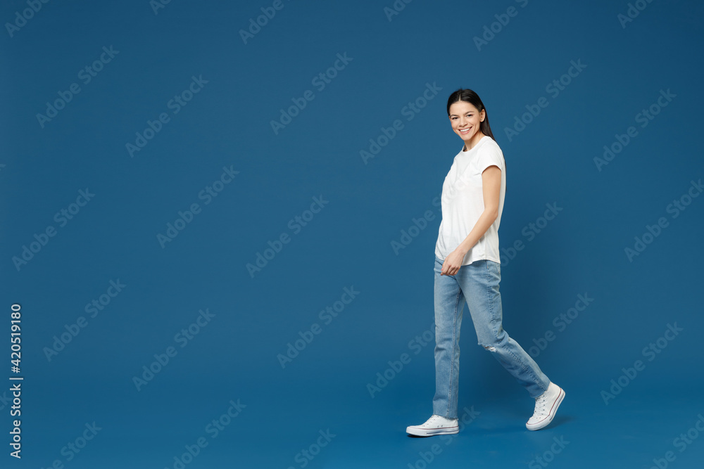 Full length side view of young smiling confident nice attractive beautiful latin woman 20s wear white casual basic t-shirt walking going look camera isolated on dark blue background studio portrait