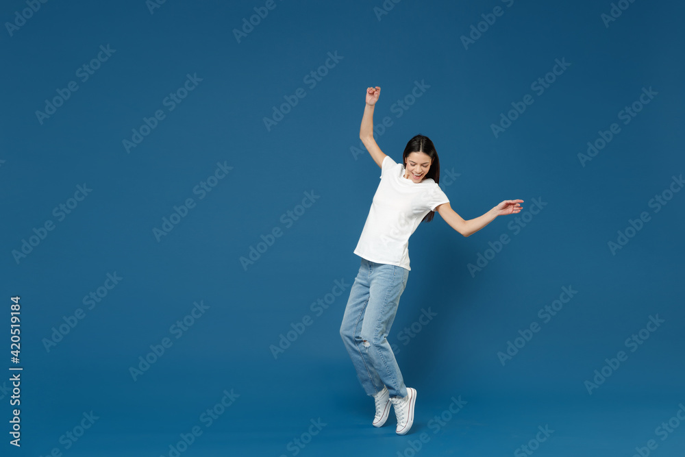 Full body length young latin woman 20s in white casual basic t-shirt keep hands outstretched leaning back look aside standing on toes dancing have fun isolated on dark blue background studio portrait.
