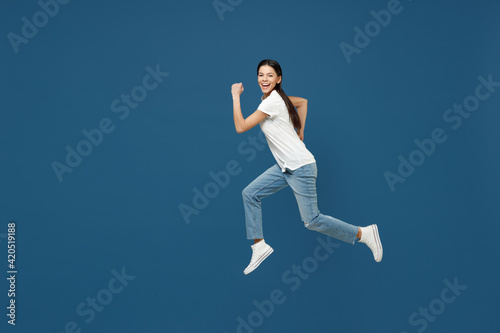 Full length side view of young happy sport smiling brunette latin woman 20s wear white casual basic t-shirt running fast hurrying jumping high isolated on dark blue color background studio portrait