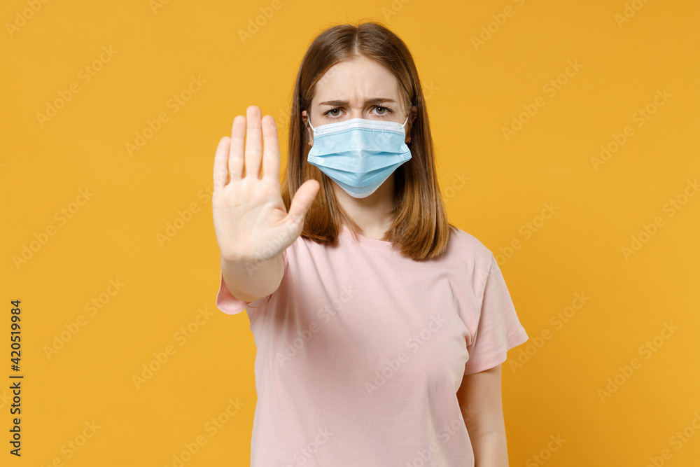 Young strict serious woman 20s in basic pastel pink t-shirt in sterile face mask, coronavirus virus covid-19 pandemic quarantine do stop palm gesture isolated on yellow background studio portrait. Young strict serious woman 20s in basic pastel pink t-shirt in sterile face mask, coronavirus virus covid-19 pandemic quarantine do stop palm gesture isolated on yellow background studio portrait.
