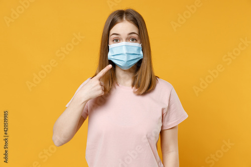 Young woman 20s in basic pastel pink t-shirt  blank print design point index finger on sterile face mask to safe from coronavirus virus covid-19  pandemic quarantine isolated on yellow background