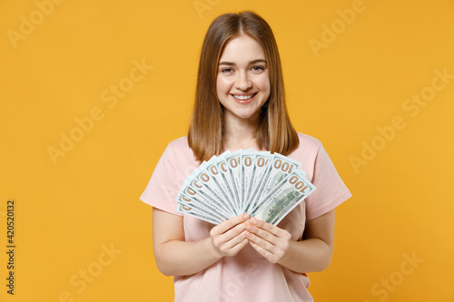 Obraz na plátně Young caucasian smiling happy rich student woman 20s wearing casual basic pastel pink t-shirt holding fan of cash money in dollar banknotes look camera isolated on yellow background studio portrait