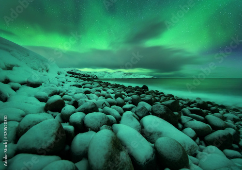 Aurora borealis over ocean. Northern lights in Teriberka, Russia. Starry sky with polar lights and clouds. Night winter landscape with bright aurora, stars, sea, snowy stones in blurred water. Travel
