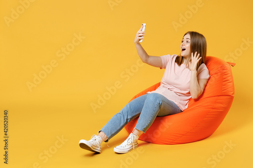 Full length of young woman 20s in basic pastel pink t-shirt, jeans sit in orange bean bag chair do selfie shot on mobile phone show waving hand greeting isolated on yellow background studio portrait.