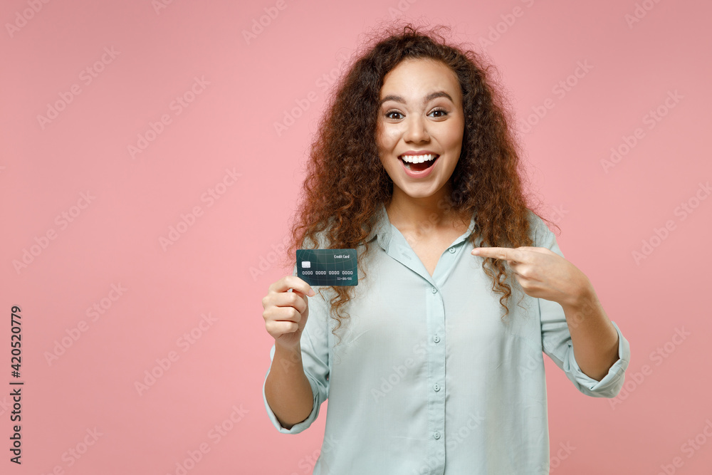 Young black african american surprised fun happy nice rich successful curly woman 20s in blue shirt pointing index finger on credit bank card isolated on pastel pink color background studio portrait Young black african american surprised fun happy nice rich successful curly woman 20s in blue shirt pointing index finger on credit bank card isolated on pastel pink color background studio portrait