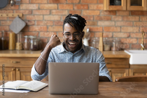 Excited black caribbean guy student laugh aloud raise winner fist see high grade for home work good exam result on laptop screen. Joyful young african man freelancer get profitable proposal by email photo
