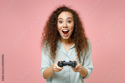 Young black african american fun surprsied happy curly female gamer woman 20s wearing casual blue shirt play pc game with joystick console isolated on pastel pink color background studio portrait. photo