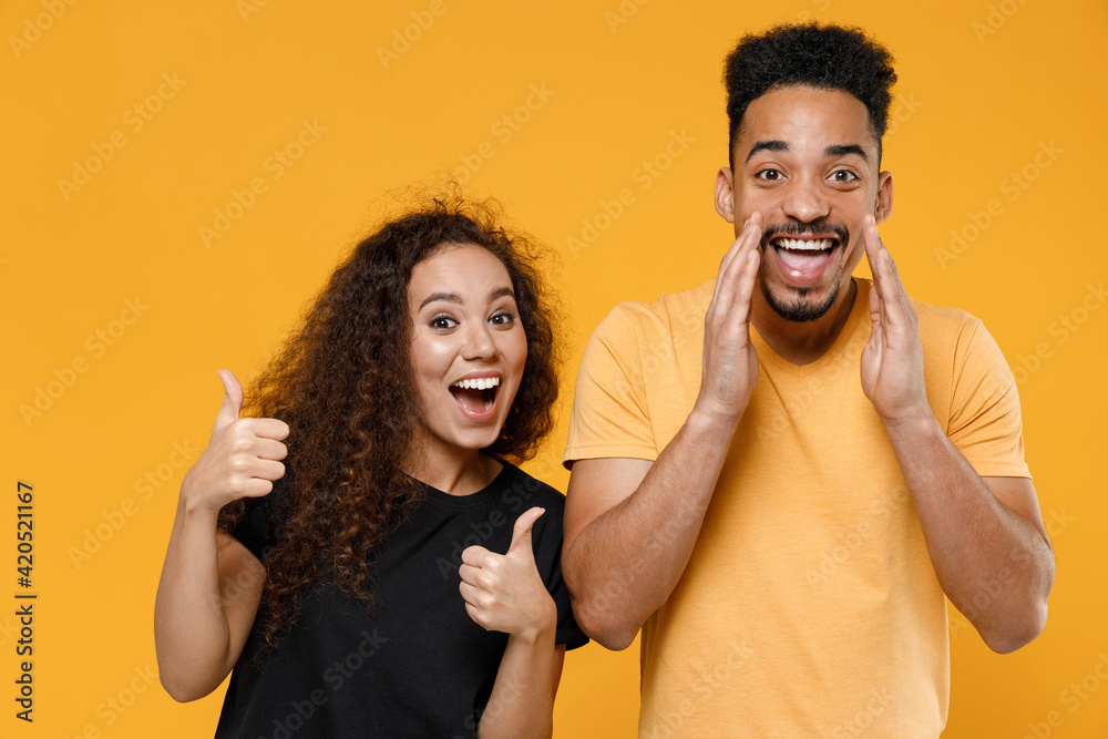 Young couple two american friends together family man african woman 20s in yellow black t-shirt show thumb up gesture scream news with hands near mouth isolated on orange background studio portrait. Young couple two american friends together family man african woman 20s in yellow black t-shirt show thumb up gesture scream news with hands near mouth isolated on orange background studio portrait.