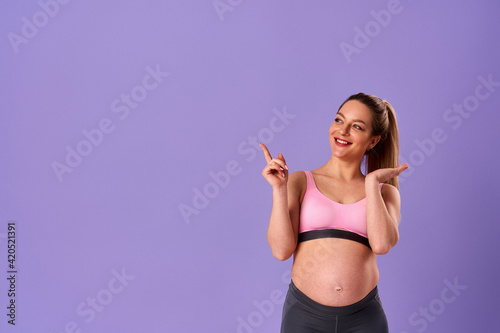 young beautiful pregnant woman, wearing sportswear on an isolated purple background, smiling, pointing her finger to the side. The expectant mother is very happy and demonstrates the copy space