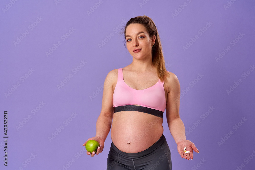 pregnant athletic woman holds pills in one hand, green apple in other, standing on purple background. pregnant blonde makes choice about which vitamins to take. Healthy pregnancy, support. pregnant athletic woman holds pills in one hand, green apple in other, standing on purple background. pregnant blonde makes choice about which vitamins to take. Healthy pregnancy, support.