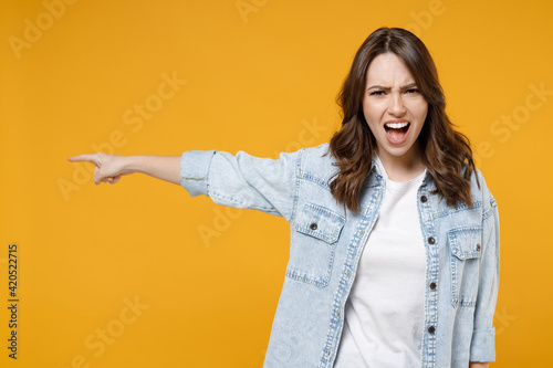 Young strict serious sad caucasian brunette woman 20s in casual stylish denim shirt white t-shirt point index finger aside commanding scream shout isolated on yellow color background studio portrait