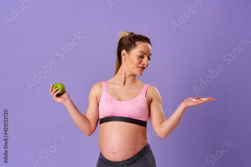 pregnant athletic woman holds pills in one hand, green apple in other, standing on purple background. pregnant blonde makes choice about which vitamins to take. Healthy pregnancy, support.