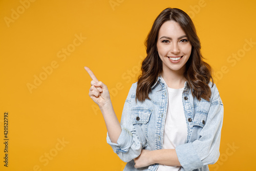 Young smiling woman promoter in denim shirt white t-shirt recommend suggest select advert point index finger aside on workspace commercial promo area mock up copy space isolated on yellow background.