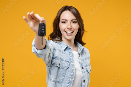 Young smiling happy satisfied excited successful brunette woman 20s in denim shirt white t-shirt hold in hands giving car keys show thumb up like gesture isolated on yellow background studio portrait.