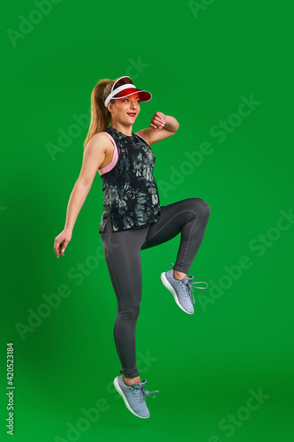 pregnant athletic woman in sportswear and a visor jumps on a green background. A pregnant blonde is engaged in sports. Healthy pregnancy, support. Sports during pregnancy. Close up, copy space.