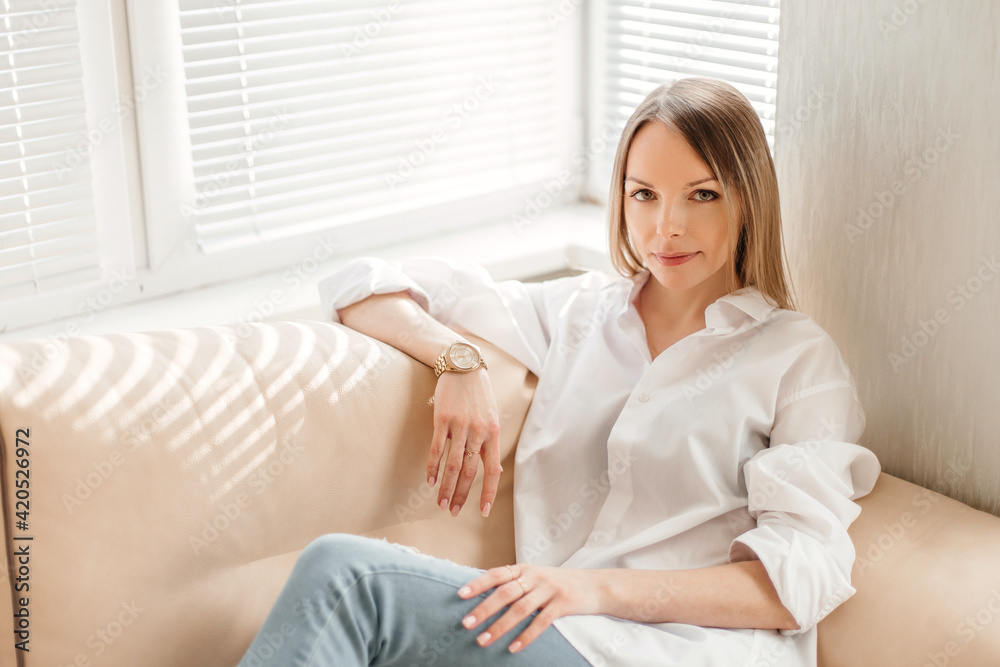 A beautiful blonde girl in a white oversized shirt and blue jeans sits in a bright sunny room on a beige sofa A beautiful blonde girl in a white oversized shirt and blue jeans sits in a bright sunny room on a beige sofa