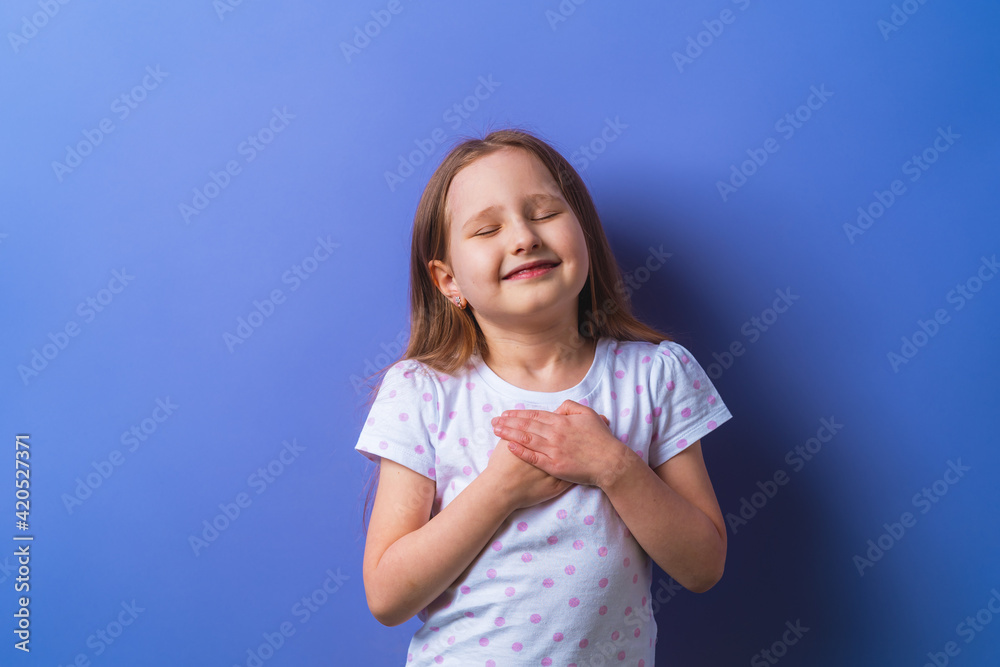 little girl with her eyes closed holds her hands on chest, feels grateful, child dreams with friendly expression, pressing her palms to her chest. concept of love and faith on a purple background