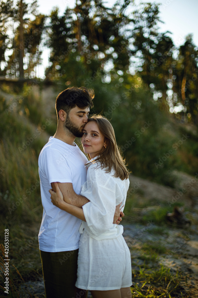 young bearded man gently kisses a young woman in white clothes in a park or forest at sunset. couple in love romantic relationship. love story