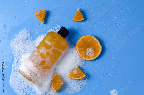Orange cosmetic product in a transparent bottle on a blue background in soapy foam with oranges. Natural cosmetic product concept copy space flat lay