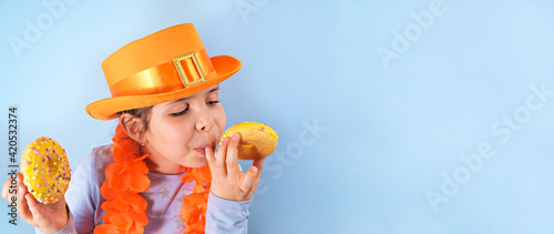 King's Day in Holland. Traditional festival on April 27 in the Netherlands. A little girl in a festive orange hat on a blue background eats colored donuts. Banner, long format. Copy space
