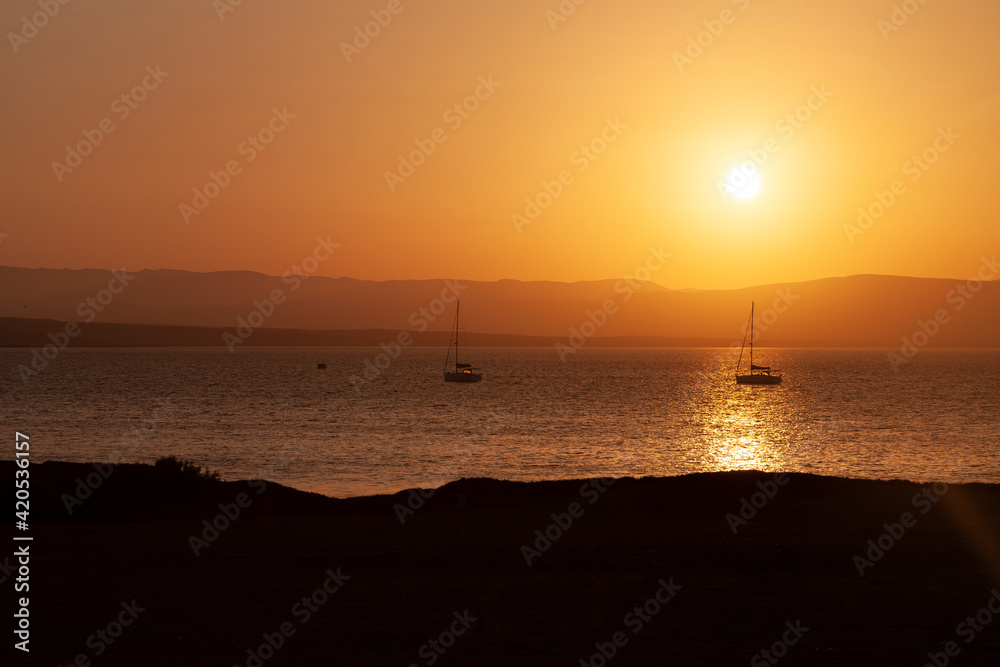 Landscape photograph at sunset, of the Paracas bay, with the silhouette of two sailboats, in the distance, outlined against the orange light, in the Paracas National Reserve, Pisco, Ica, Peru.