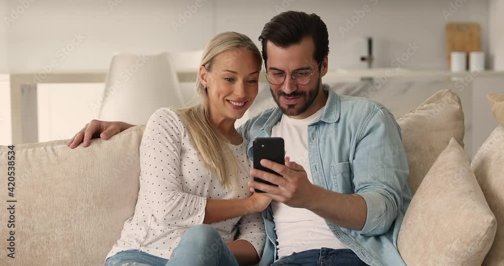 Millennial couple rest on sofa having fun using smartphone mobile cool app. Spouses discussing online purchase in e-commerce retail web sites, make order, buying goods and services on internet concept Millennial couple rest on sofa having fun using smartphone mobile cool app. Spouses discussing online purchase in e-commerce retail web sites, make order, buying goods and services on internet concept