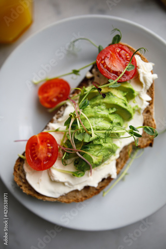 avocado toast from whole grain bread with cream cheese tomatoes and microgreen. sandwich on a plate on a white marble surface. vegan breakfast