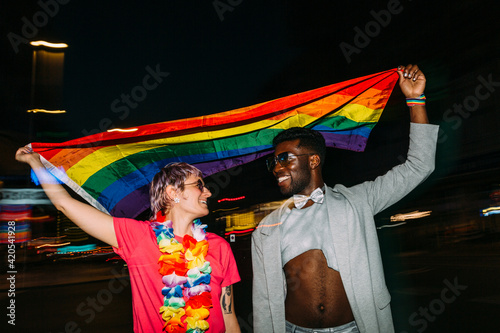 Happy multiracial couple raising LGBT pride flag in street at ni photo