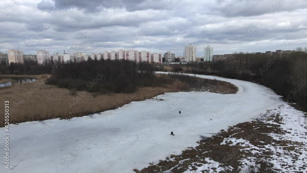 Flying over a river covered with ice. Fishermen are sitting on the ice. Winter fishing. Multi-storey buildings are visible on the horizon. Flying over a river covered with ice. Fishermen are sitting on the ice. Winter fishing. Multi-storey buildings are visible on the horizon.